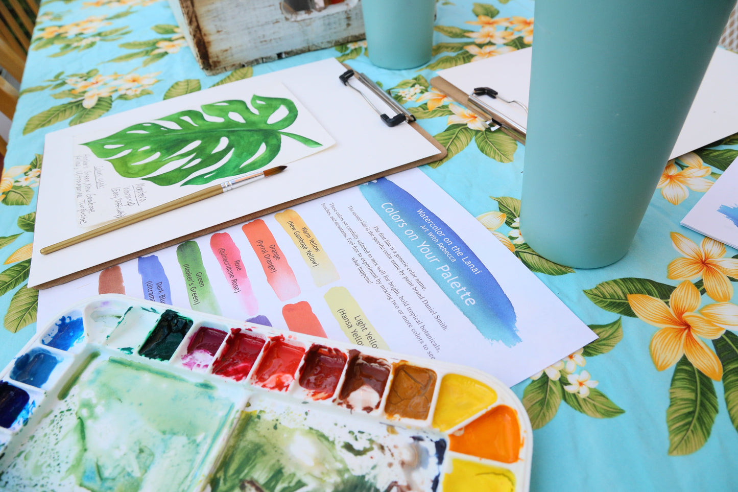 Art supplies including a watercolor palette, brushes, and a color chart on a floral tablecloth.