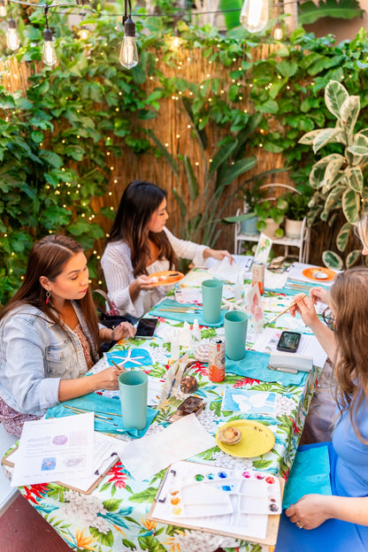 Three women painting watercolor at a table outdoors with plants and string lights in the background.